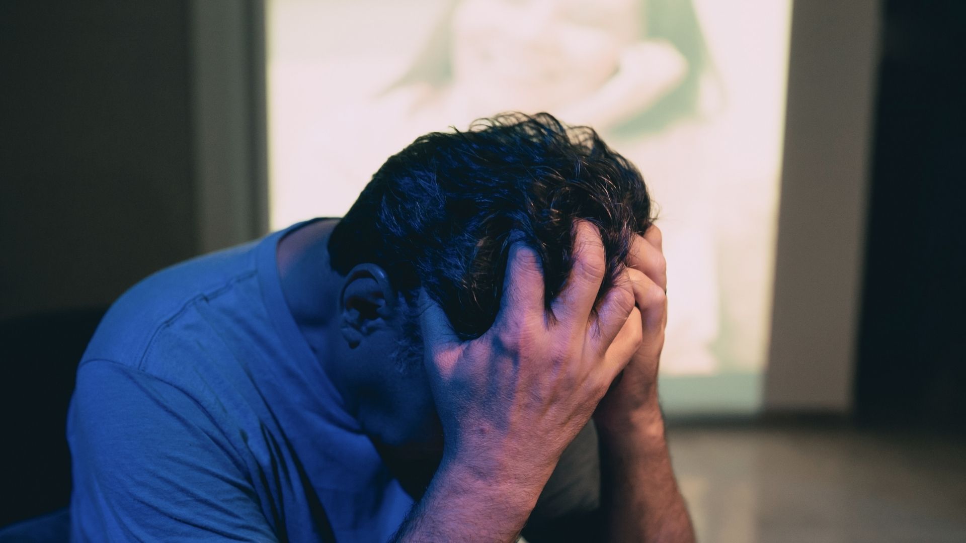 Man with head in hands, appearing distressed, in a dimly lit room.